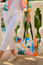 A person wearing white pants and a colorful tropical print jacket holds a large tote bag with a matching tropical architectural print. The bag features black rope handles and is set against a white balustrade with green foliage in the background.