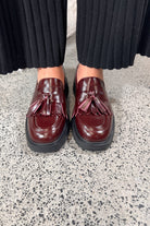Person wearing maroon loafers with tassels on a speckled gray floor.