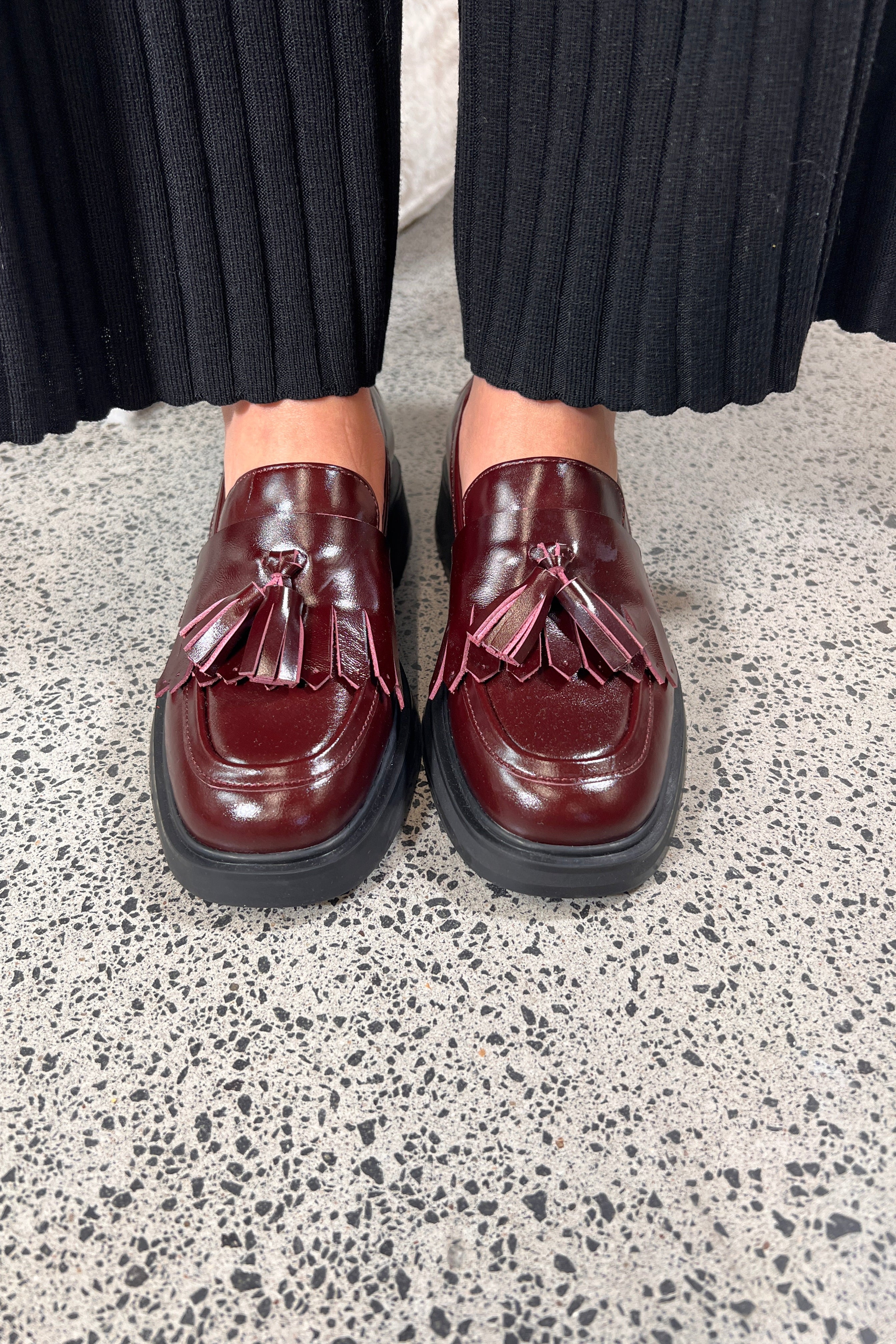Person wearing maroon loafers with tassels on a speckled gray floor.