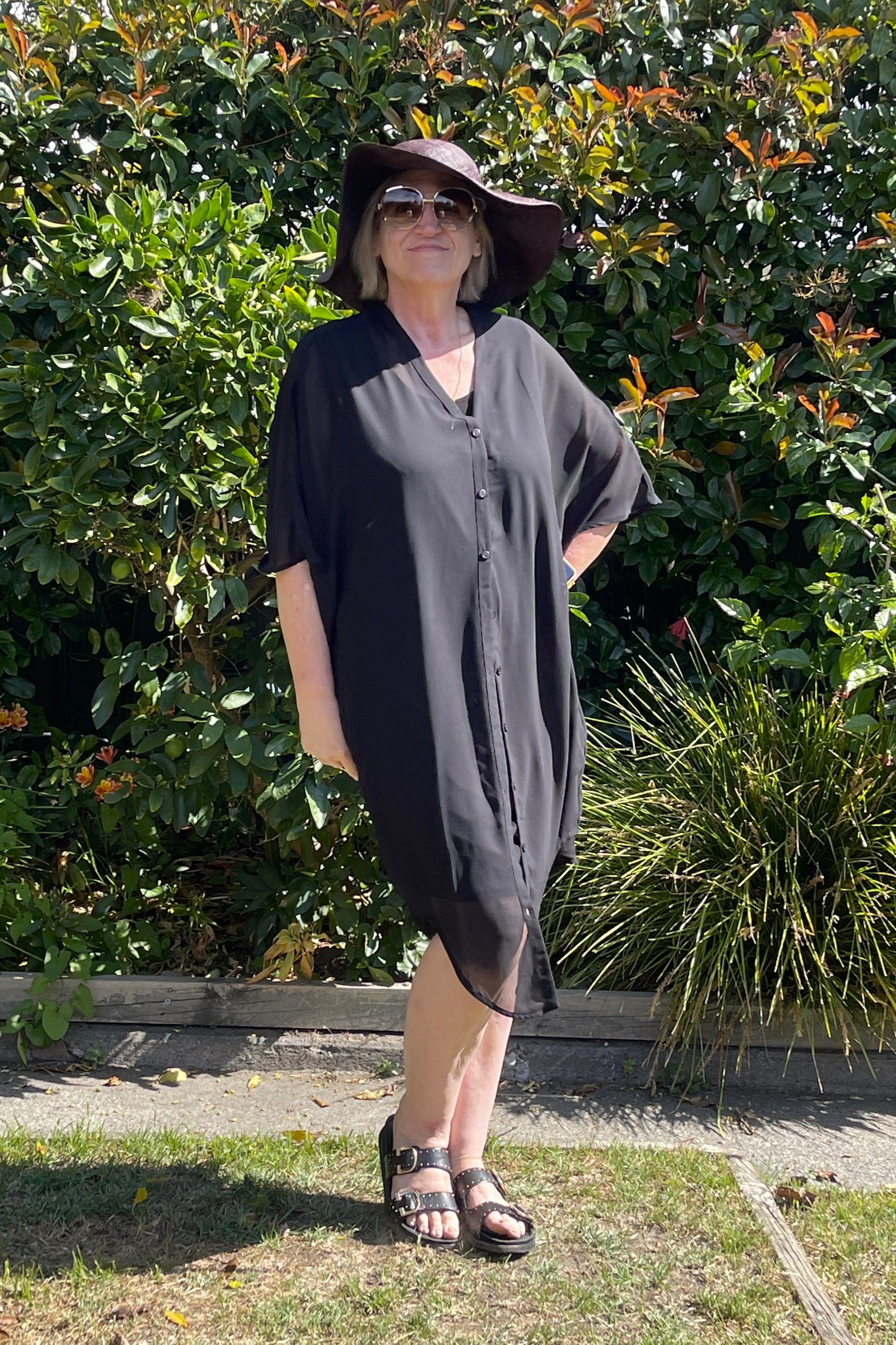 Woman wearing a black kaftan and hat standing in front of green bushes