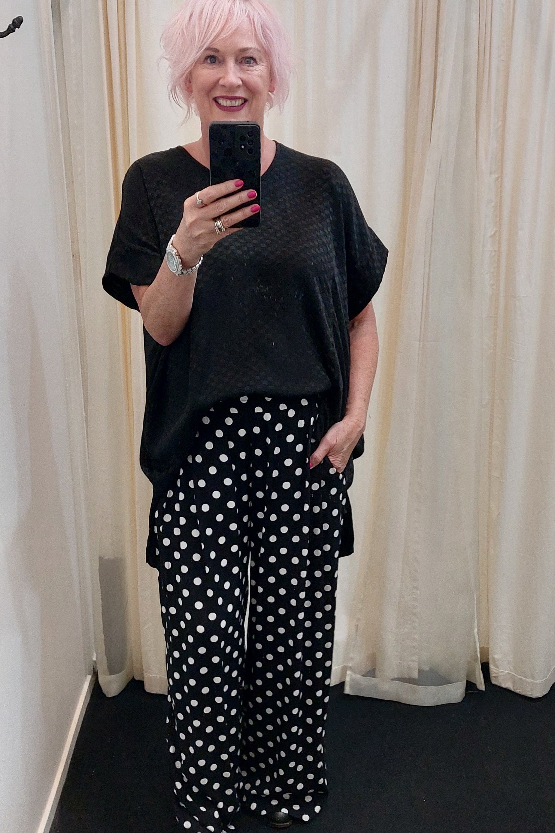 Person taking a mirror selfie wearing a black top and polka dot pants in front of white curtains.