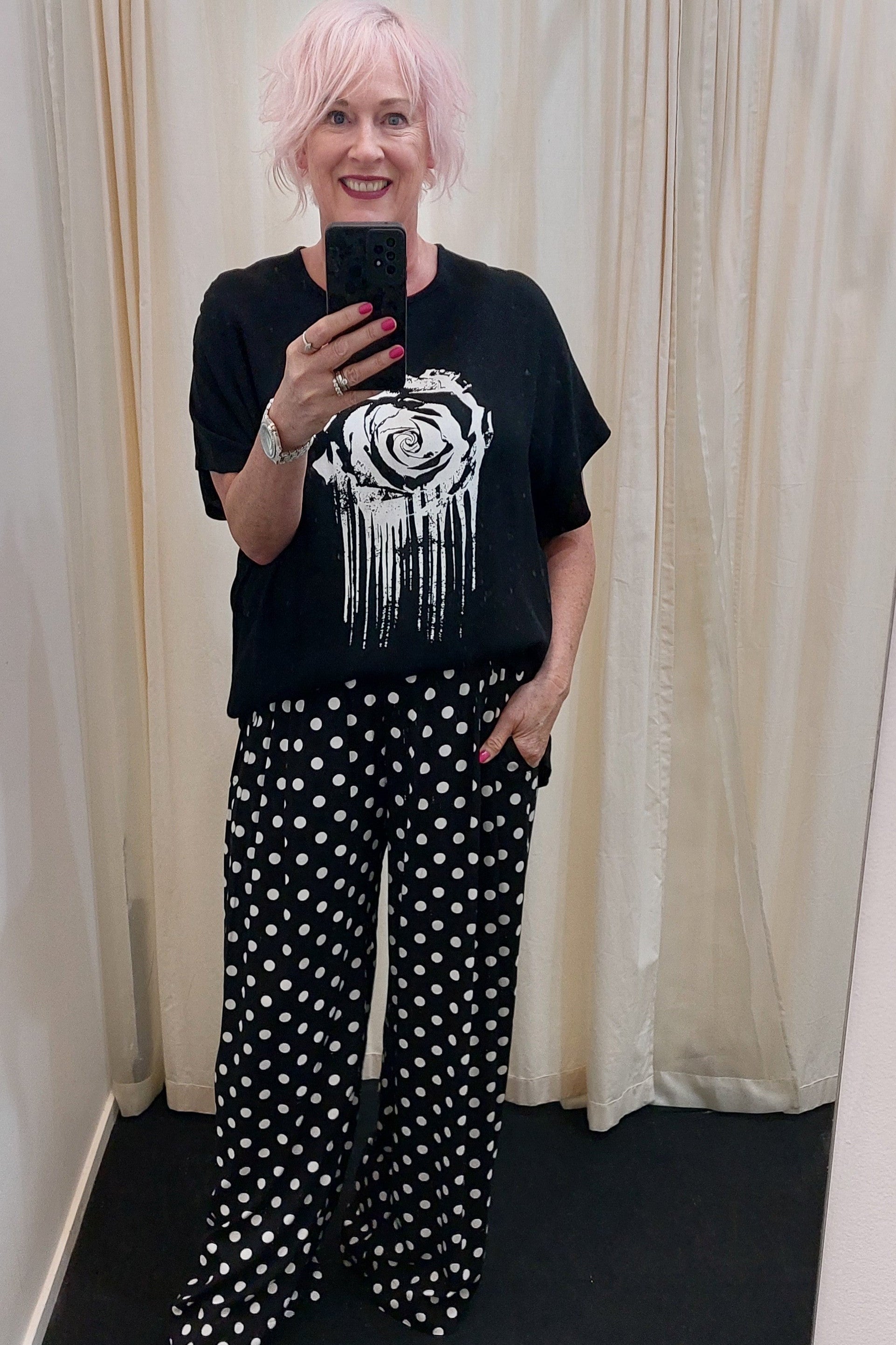 Person wearing a black t-shirt with a graphic design and polka dot pants, standing against a white curtain.