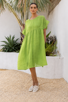 A woman wearing a bright lime green short-sleeve midi dress and white patterned flat shoes, standing in a sunny outdoor courtyard with a white wall, plants, and gravel.