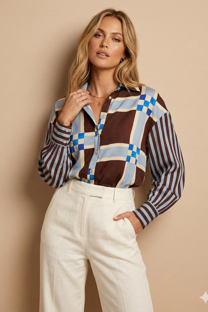 A woman models a long-sleeve button-up shirt featuring a brown, blue, and beige geometric grid pattern with striped sleeves, paired with cream-colored trousers, set against a solid beige background.