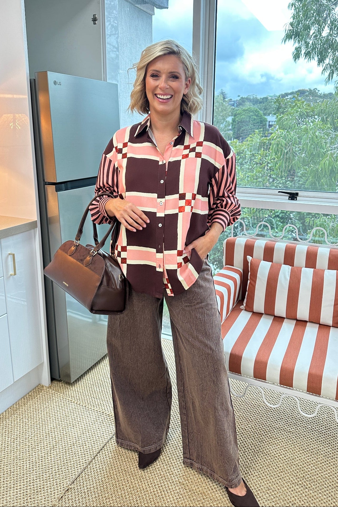 A woman stands in a modern kitchen and seating area wearing a brown, pink, and white plaid button-down shirt, wide-leg brown pants, and holding a brown handbag.