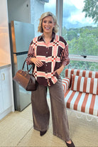 A woman stands in a modern kitchen and seating area wearing a brown, pink, and white plaid button-down shirt, wide-leg brown pants, and holding a brown handbag.