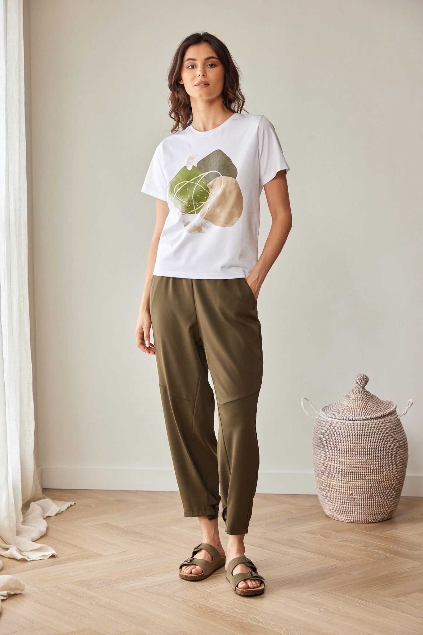 A woman stands in a bright room wearing a white graphic T-shirt with a green and beige fruit print, olive green cropped pants, and brown slide sandals. A woven basket and light wood floor are visible in the background.