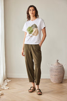 A woman stands in a bright room wearing a white graphic T-shirt with a green and beige fruit print, olive green cropped pants, and brown slide sandals. A woven basket and light wood floor are visible in the background.