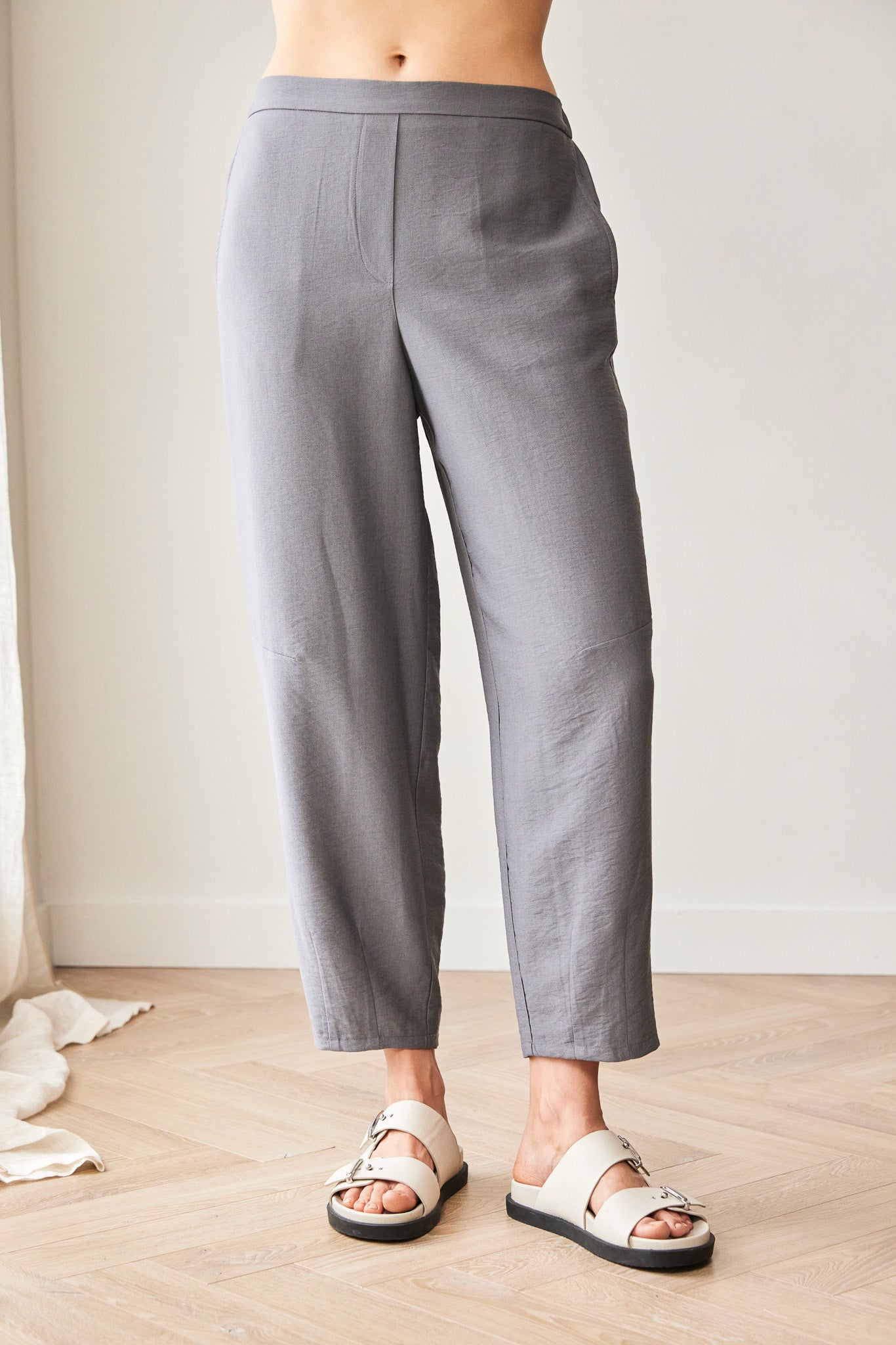 A person wearing loose, straight-leg cropped trousers in a medium gray color, paired with off-white slide sandals, standing on a light wood floor against a neutral wall.