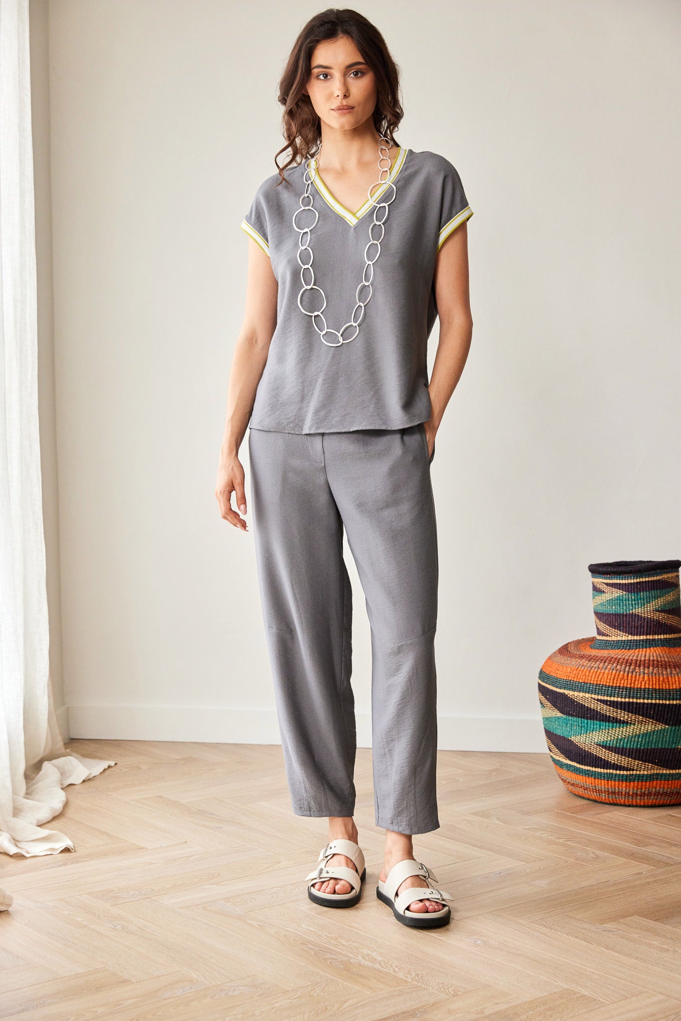 A woman stands in a bright indoor room wearing a gray short-sleeve V-neck top with yellow trim and matching gray cropped pants, accessorized with a chunky silver necklace and white slide sandals.