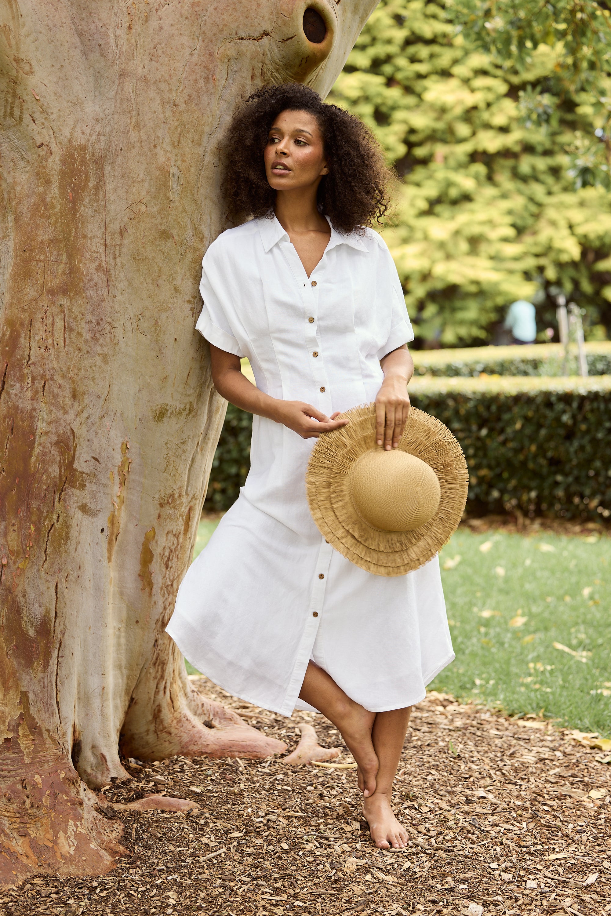 Woman in a white dress holding a straw hat, standing next to a tree in an outdoor setting.
