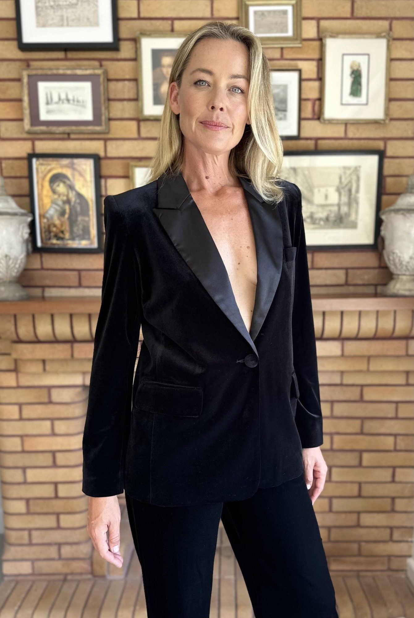 Person wearing a black suit with a deep neckline against a wooden wall with framed pictures.