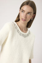 Woman wearing a white sweater with a decorative neckline against a plain background