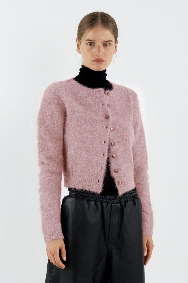 Person wearing a pink cardigan over a black turtleneck and pants on a white background