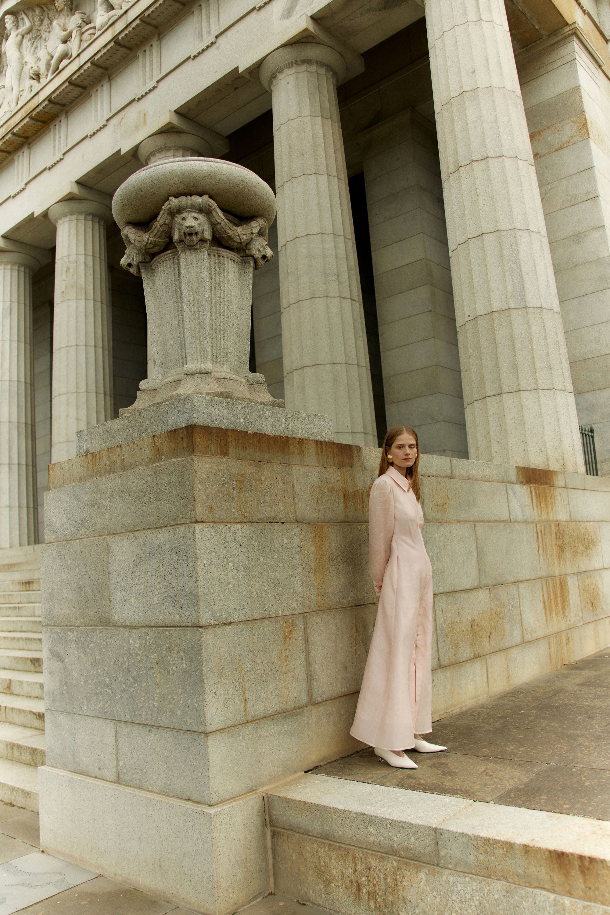Woman in a long light pink dress standing on stone steps in front of classical architecture.