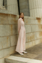 Woman in a long light pink dress standing on stone steps against a stone wall.
