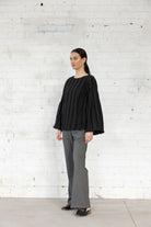 Woman wearing a black top and gray pants standing against a white brick wall.