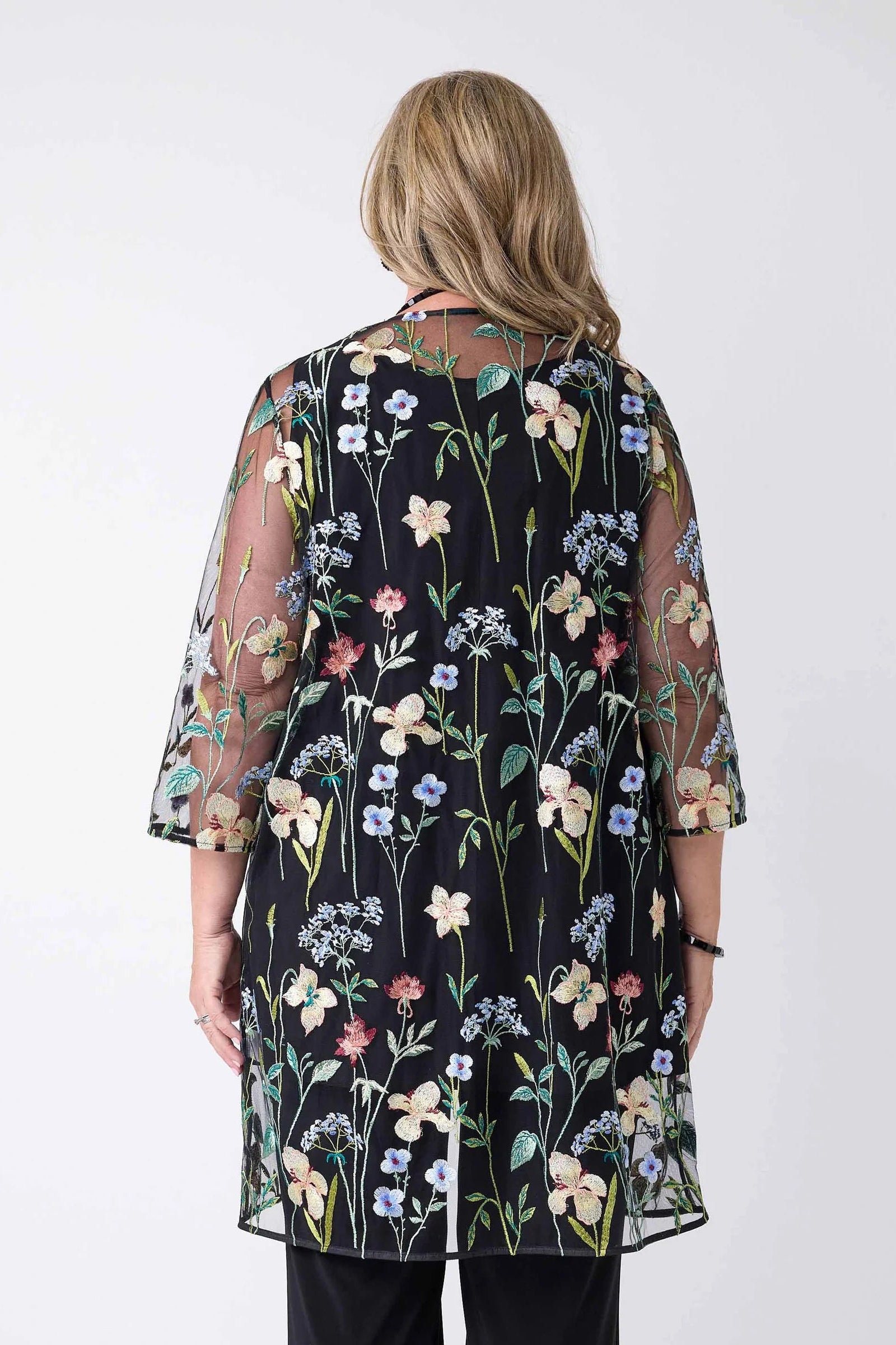 A woman viewed from the back wearing a sheer black tunic-style top with a colorful floral print and elbow-length sleeves, paired with black pants, against a plain light gray studio background.