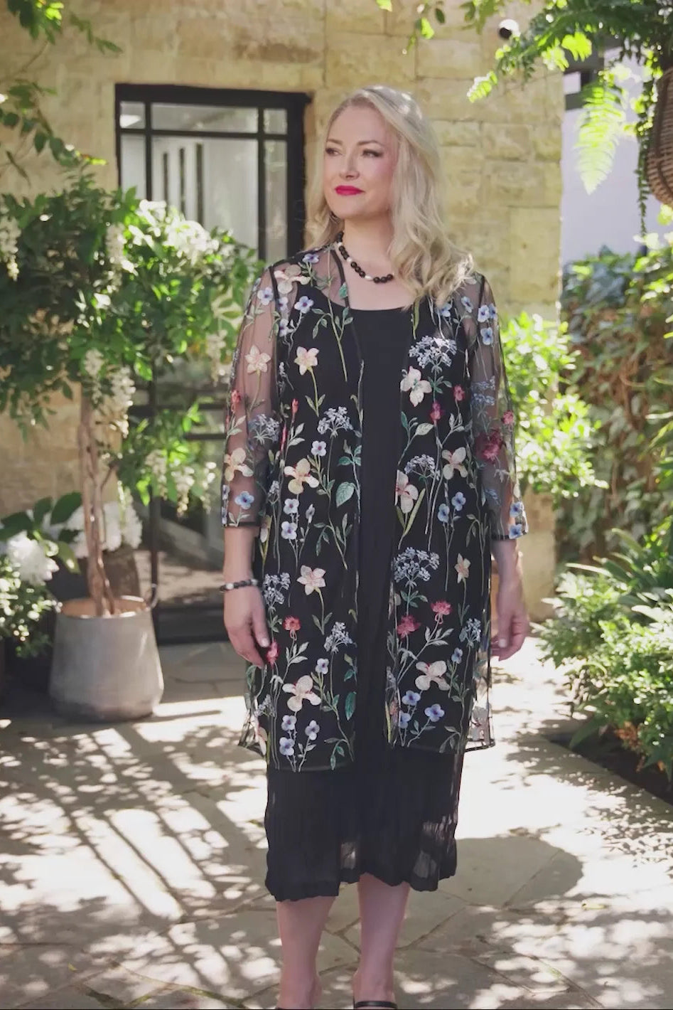 A woman stands in a sunlit garden courtyard wearing a sheer black floral cardigan over a black dress. The cardigan features embroidered flowers in pink, blue, white, and yellow with green stems.