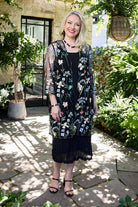 A woman stands outdoors in a garden courtyard wearing a black sheer embroidered tunic dress with floral patterns in white, blue, and green. She is wearing black strappy high-heeled sandals and a black necklace.