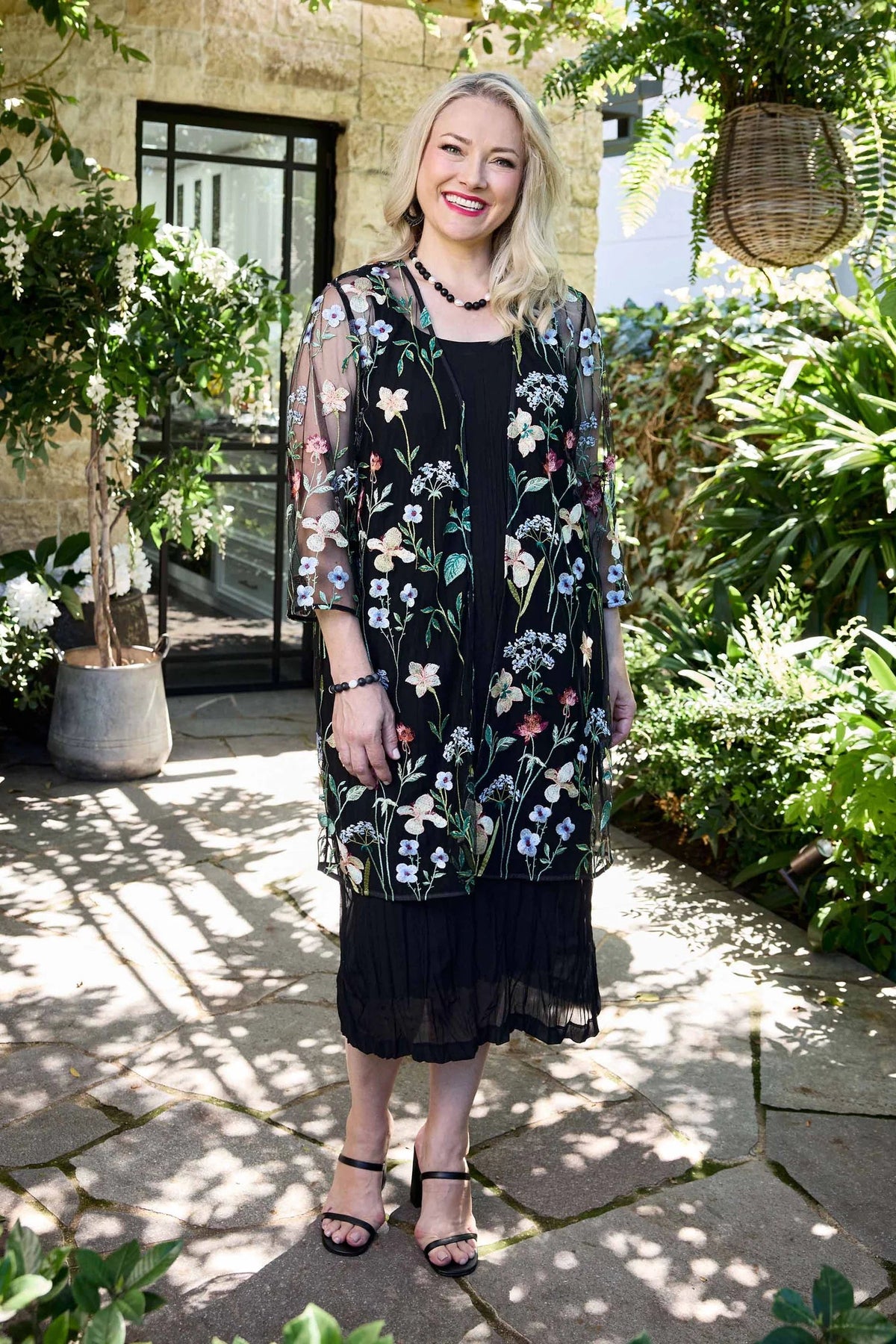 A woman stands outdoors in a garden courtyard wearing a black sheer embroidered tunic dress with floral patterns in white, blue, and green. She is wearing black strappy high-heeled sandals and a black necklace.