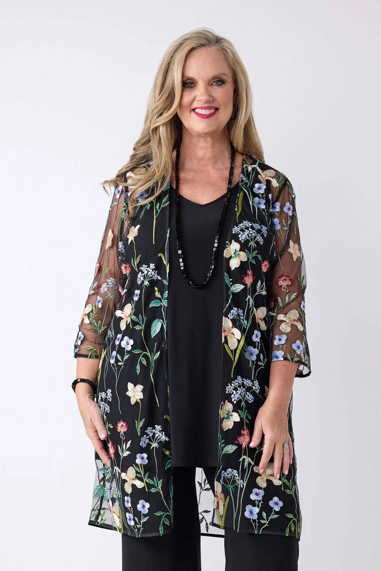 A woman modeling a sheer black floral kimono cardigan with three-quarter sleeves over a black top, photographed against a plain light gray studio background.