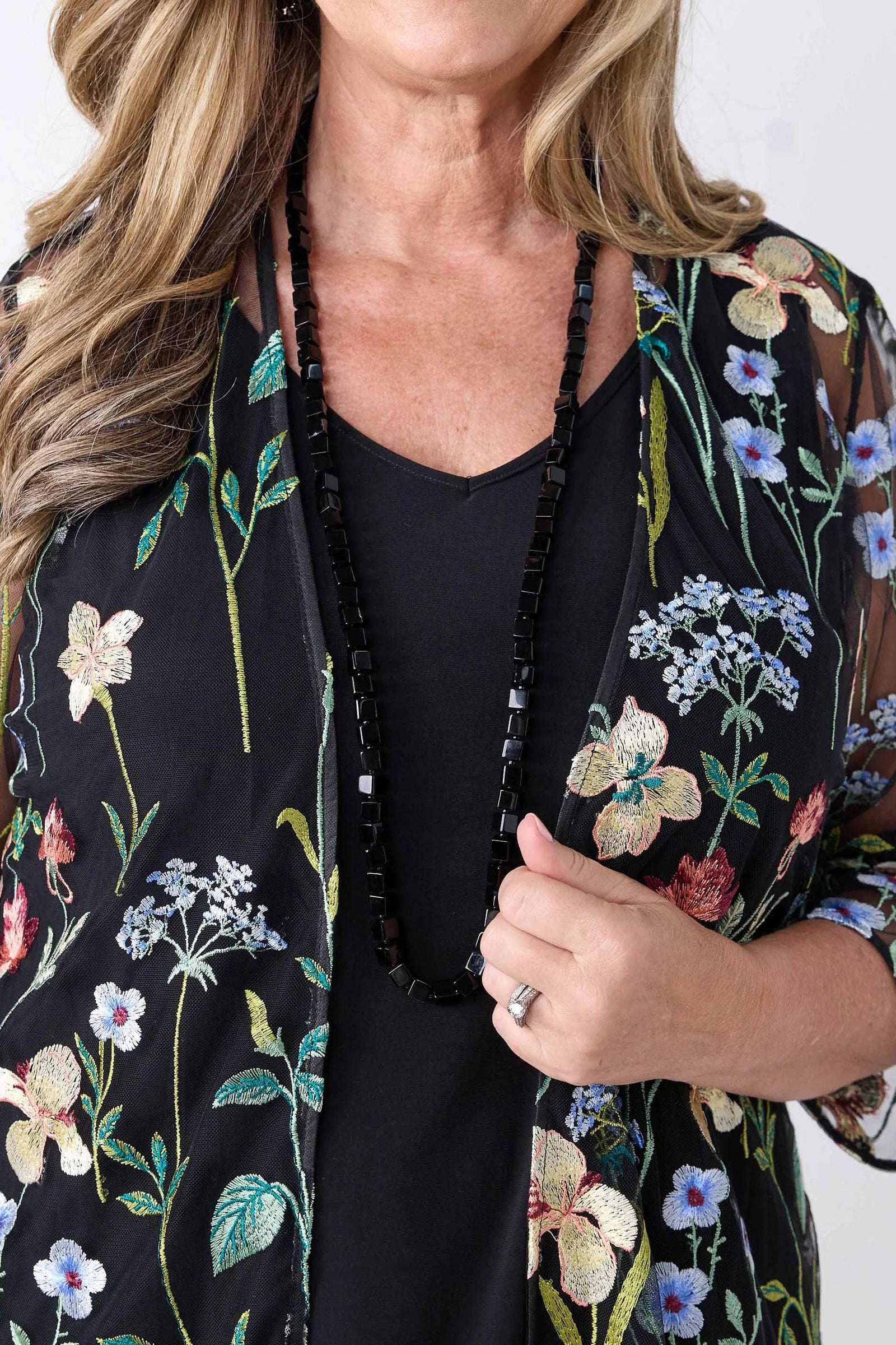 A close-up of a woman wearing a black sheer floral kimono cardigan with embroidered flowers and green leaves over a black top, accessorized with a long black beaded necklace.