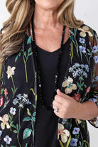 A close-up of a woman wearing a black sheer floral kimono cardigan with embroidered flowers and green leaves over a black top, accessorized with a long black beaded necklace.