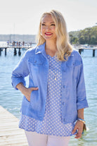 A woman standing on a wooden dock by the water wearing a light blue denim-style jacket over a white and blue polka dot top and white pants.