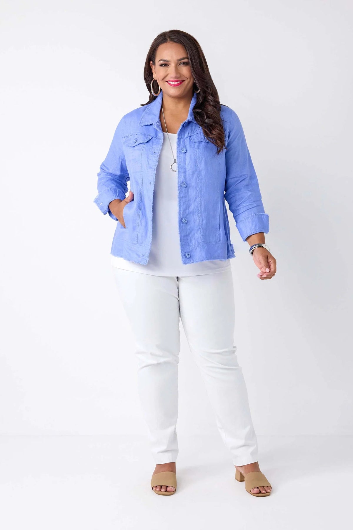 A woman modeling a light blue button-up shirt jacket over a white top and white pants, standing against a plain white studio background.