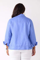 A woman viewed from the back wearing a light blue cropped jacket with rolled sleeves and white pants, against a plain white studio background.