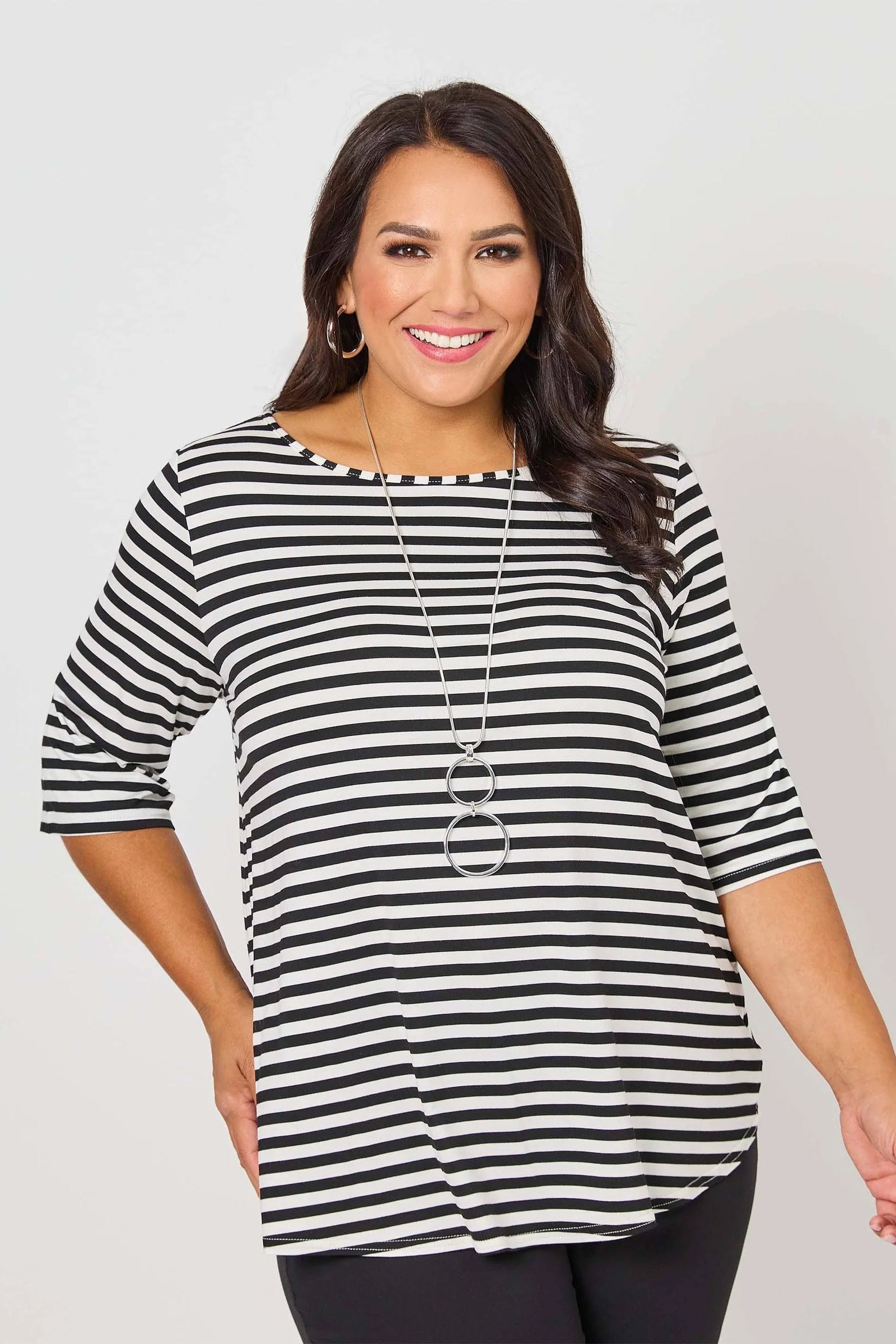 A woman modeling a black and white horizontally striped short-sleeve top with a boat neckline, paired with black pants and a long silver necklace, against a light gray studio background.