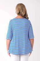 A woman viewed from the back wearing a blue and white horizontally striped short-sleeve top with white pants, set against a plain white studio background.