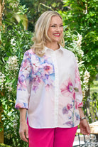 Woman wearing a floral blouse and pink pants standing outdoors with greenery in the background
