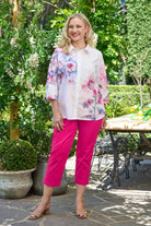 Woman wearing a floral blouse and pink pants standing outdoors in a garden setting.