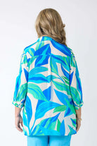 A woman viewed from behind wearing a vibrant tropical leaf print shirt with blue, green, and white leaves, paired with light blue pants, against a plain white background.
