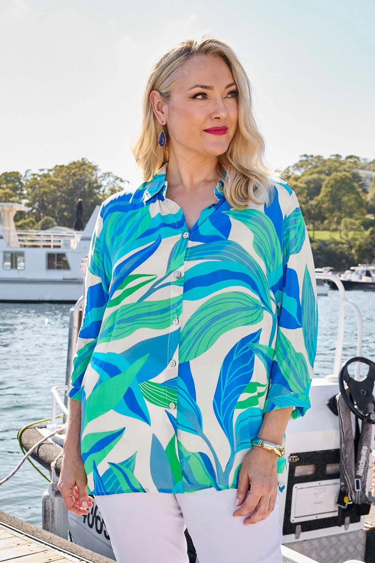 A woman wearing a vibrant blue and green tropical leaf print button-up shirt and white pants, standing on a boat deck with water and boats in the background.
