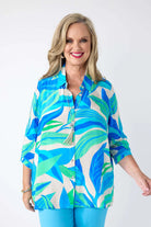 A smiling blonde woman wearing a colorful tropical leaf print button-up shirt with blue and green leaves on a cream background, paired with bright blue pants, against a plain white studio background.