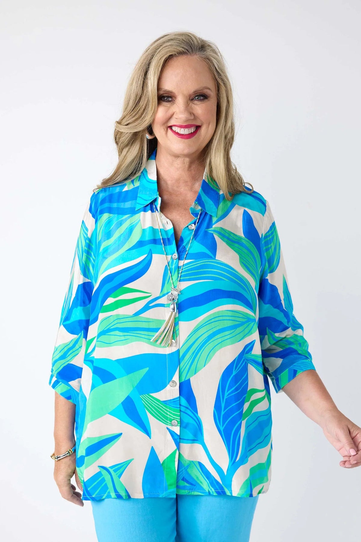A smiling blonde woman wearing a colorful tropical leaf print button-up shirt with blue and green leaves on a cream background, paired with bright blue pants, against a plain white studio background.