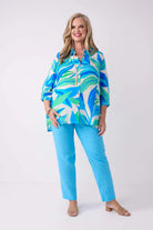 A woman modeling a vibrant tropical print button-up shirt in shades of blue, green, and white, paired with solid bright blue pants, against a clean white studio background.