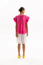 A woman shown from the back wearing a bright pink short-sleeve blouse with ruffled sleeves and white shorts, standing against a plain white studio background.