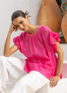 A woman wearing a bright fuchsia pink short-sleeve blouse with ruffled details and white pants, posing in front of a white wall with large terracotta pots.