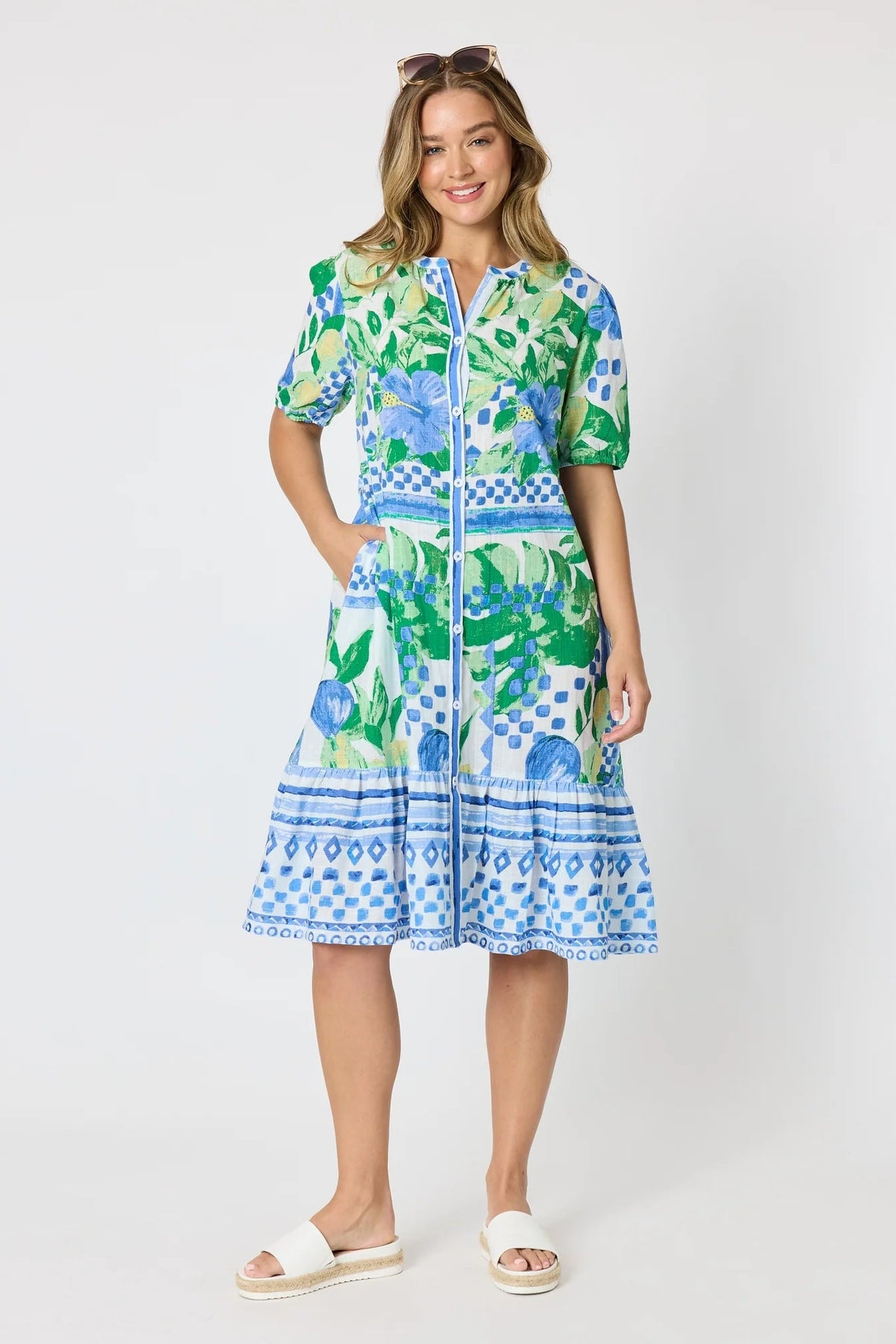 Woman wearing a button up knee length summer dress featuring Mediterranean print with botanical, lemon and blue geometric design against a white background.