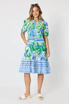 Woman wearing a button up knee length summer dress featuring Mediterranean print with botanical, lemon and blue geometric design against a white background.
