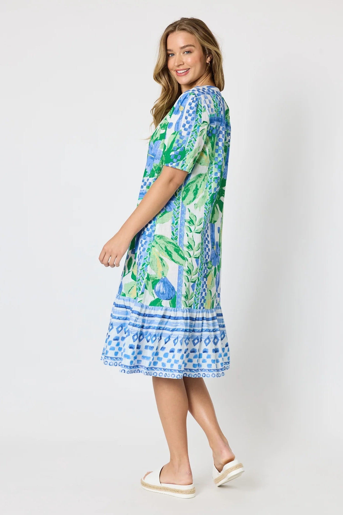 Woman wearing a button up knee length summer dress featuring Mediterranean print with botanical, lemon and blue geometric design against a white background.