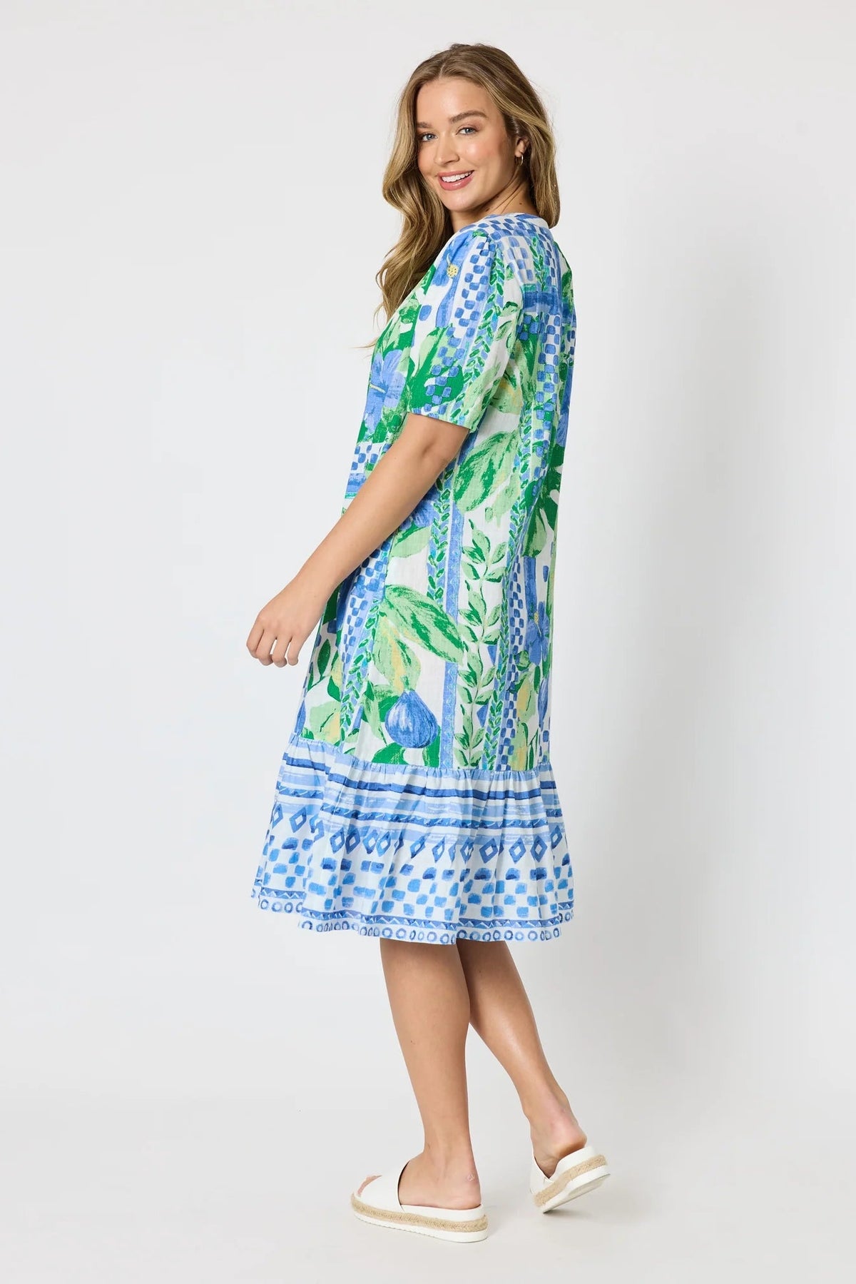 Woman wearing a button up knee length summer dress featuring Mediterranean print with botanical, lemon and blue geometric design against a white background.