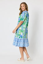 Woman wearing a button up knee length summer dress featuring Mediterranean print with botanical, lemon and blue geometric design against a white background.