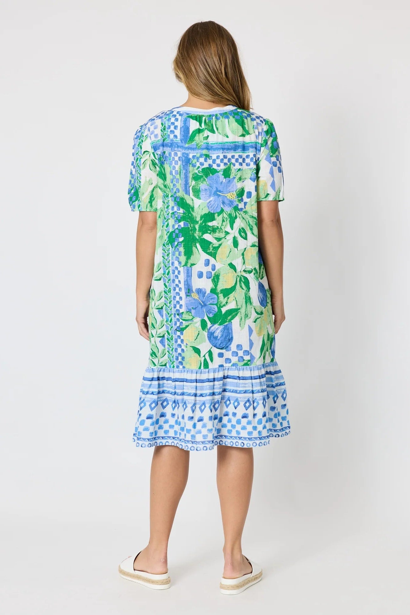 Woman wearing a button up knee length summer dress featuring Mediterranean print with botanical, lemon and blue geometric design against a white background.