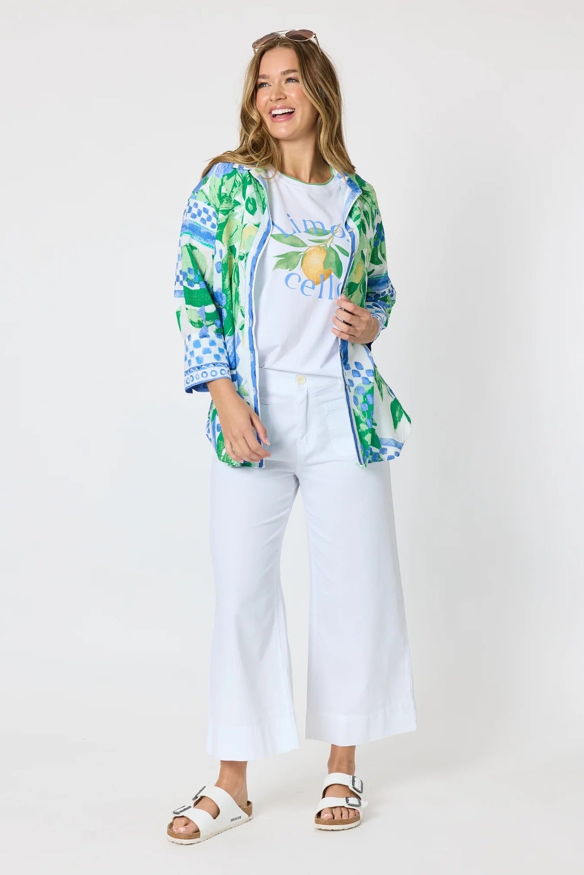 Woman wearing a button up shirt with collar and Mediterranean print with botanical, lemon and blue geometric design and white pants and white background.