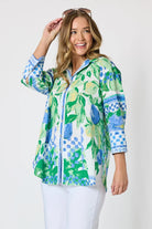 Woman wearing a button up shirt with collar and Mediterranean print with botanical, lemon and blue geometric design and white pants and white background.